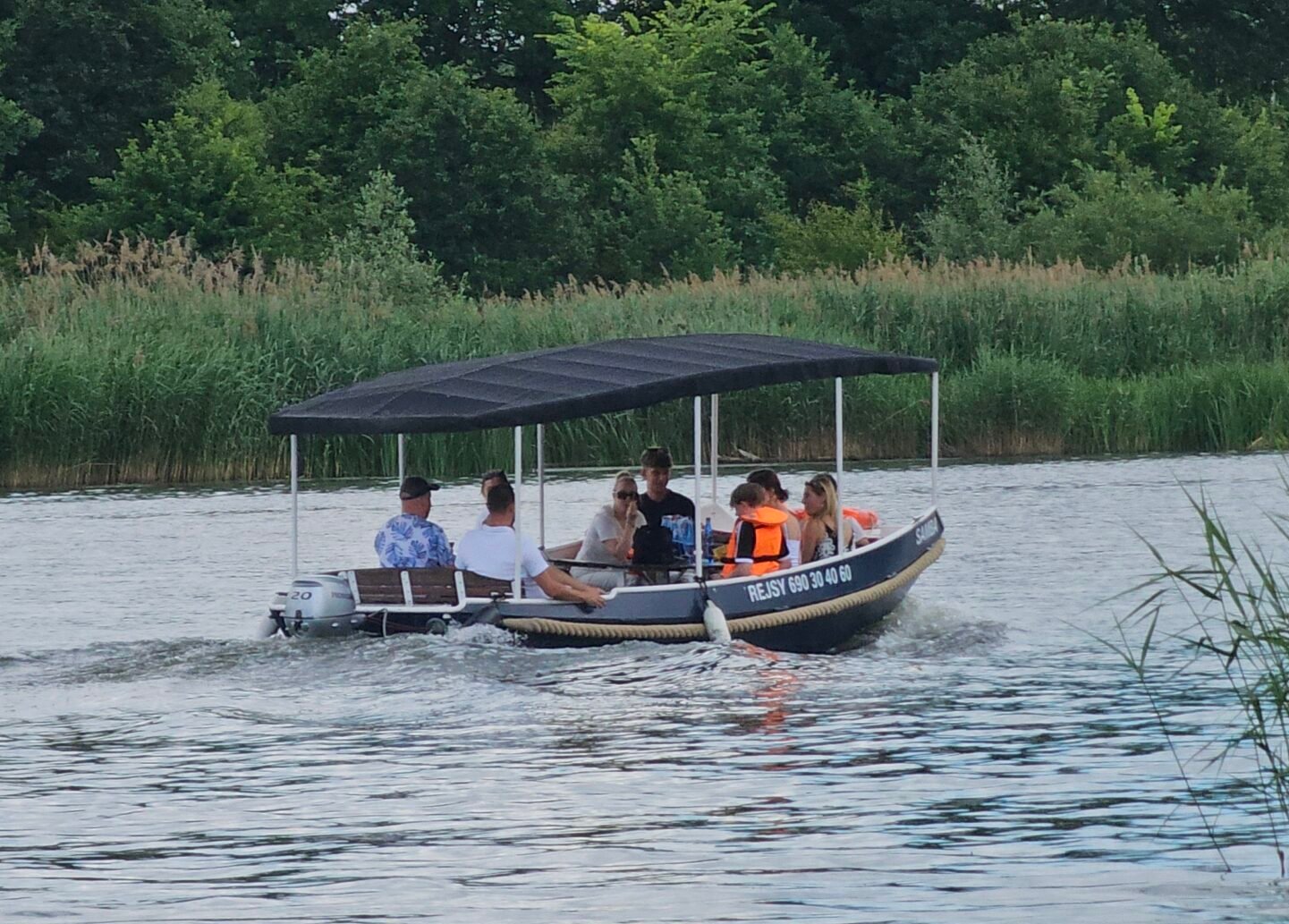 1 - portosobowice.pl A covered pontoon boat with people aboard traveling on a calm river, backed by tall reeds and trees, some wearing life jackets.