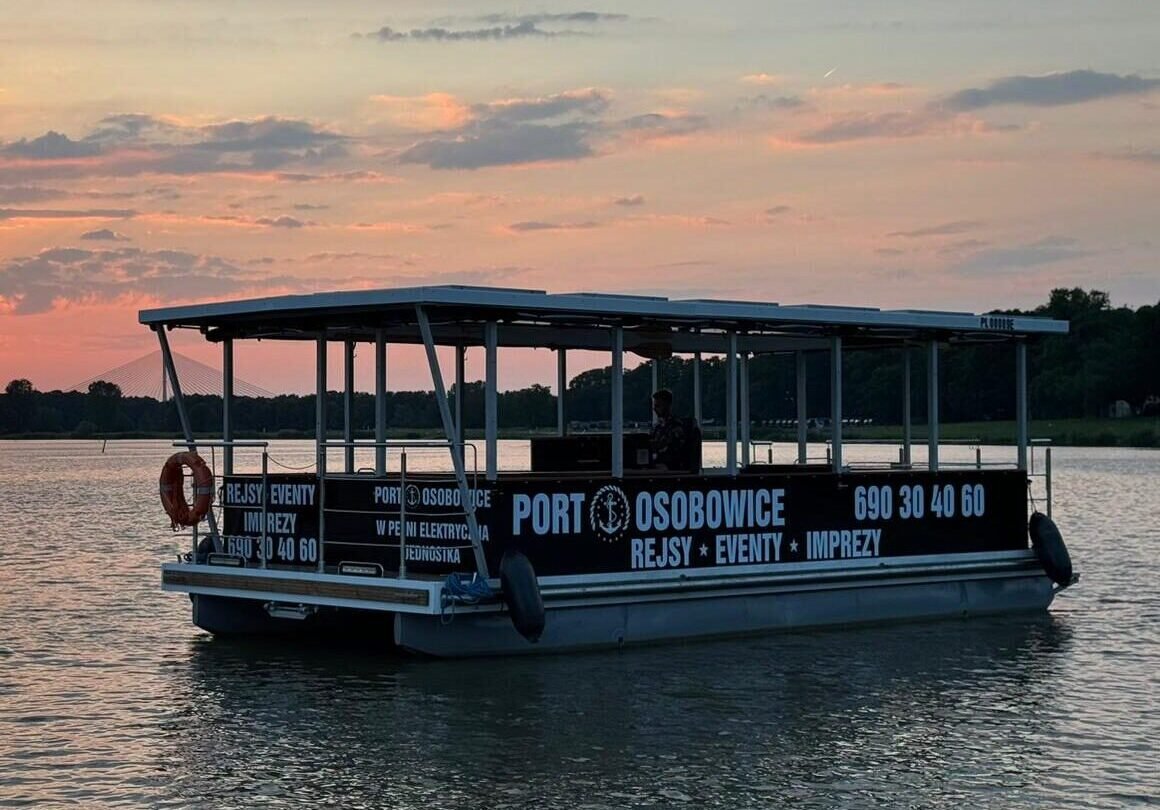 Sunset over a lake with a covered passenger boat labeled Port Osobowice, reflecting orange light on the water.