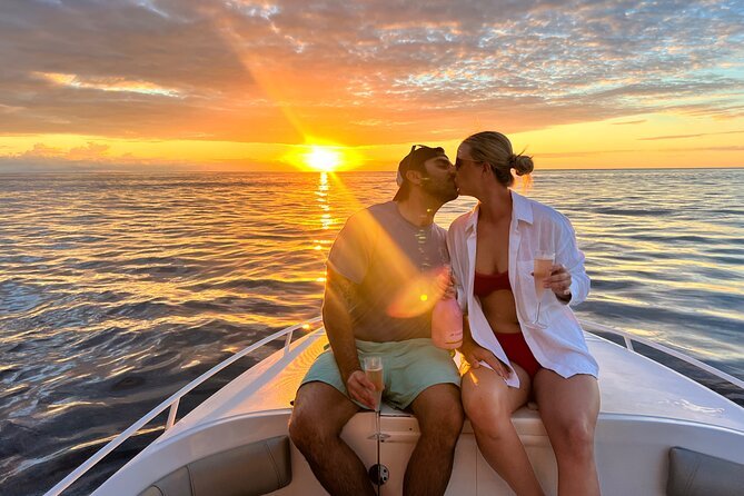 Couple kissing on the bow of a boat at sunset, each holding a drink, with the ocean and a vibrant orange sky in the background.